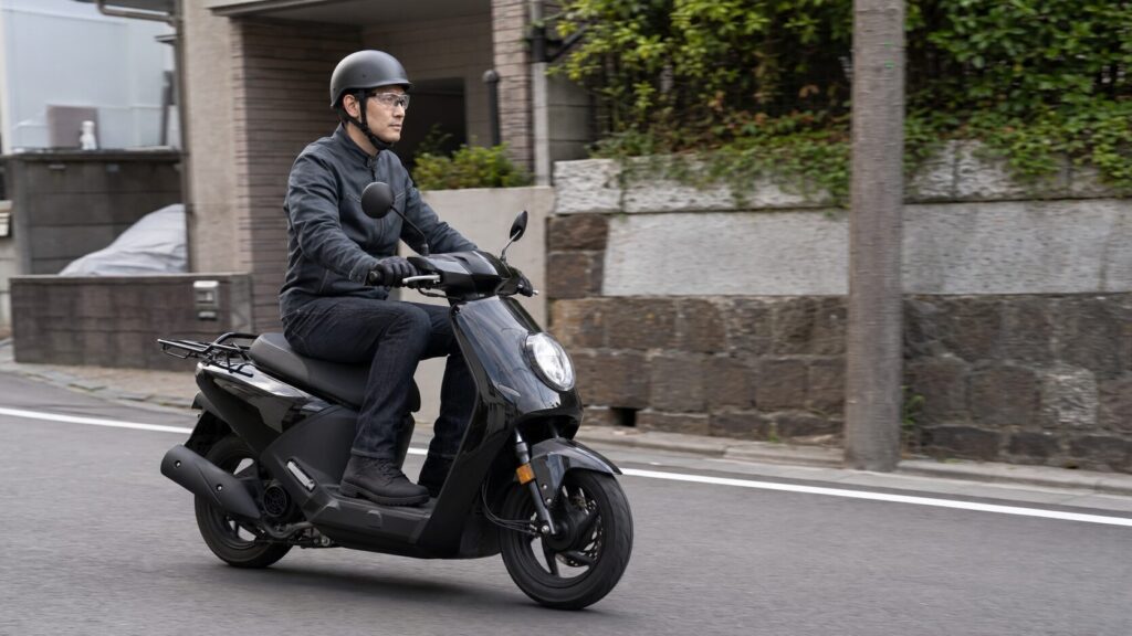 半キャップと保護メガネと防護服で街を走る日本人スクーターライダー
